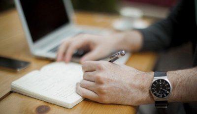 A left-handed person writes notes in their moleskin notebook